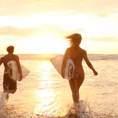 Two people holding surfboards running into the ocean as the sun rises in the distance.